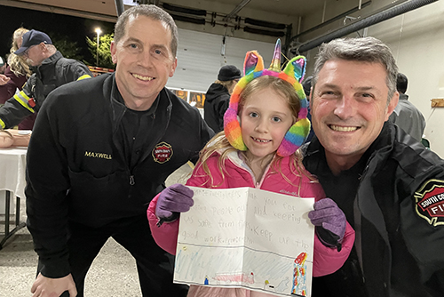 Two fire chiefs with a girl holding a thank you note to firefighters