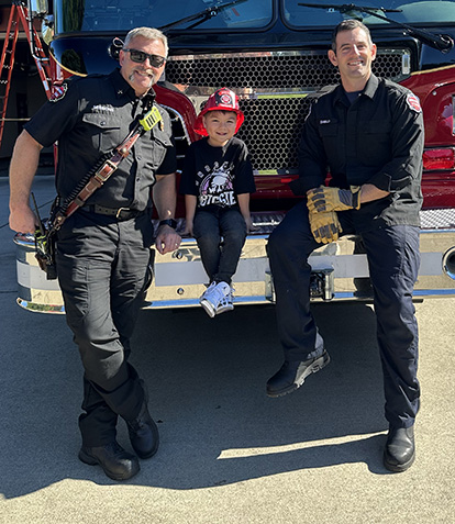 Two firefighters and a boy sitting on the front bumper of a fire engine